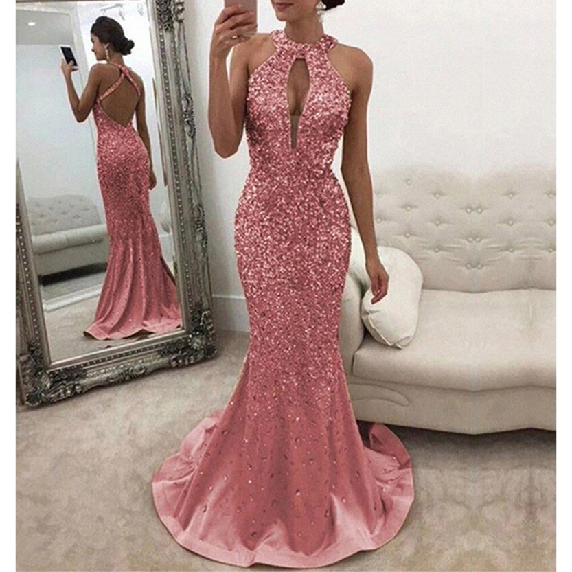 A woman wearing sparkling pink evening gown with a halter neck and open back stands by a mirror. The setting is elegant, with a plush beige sofa.